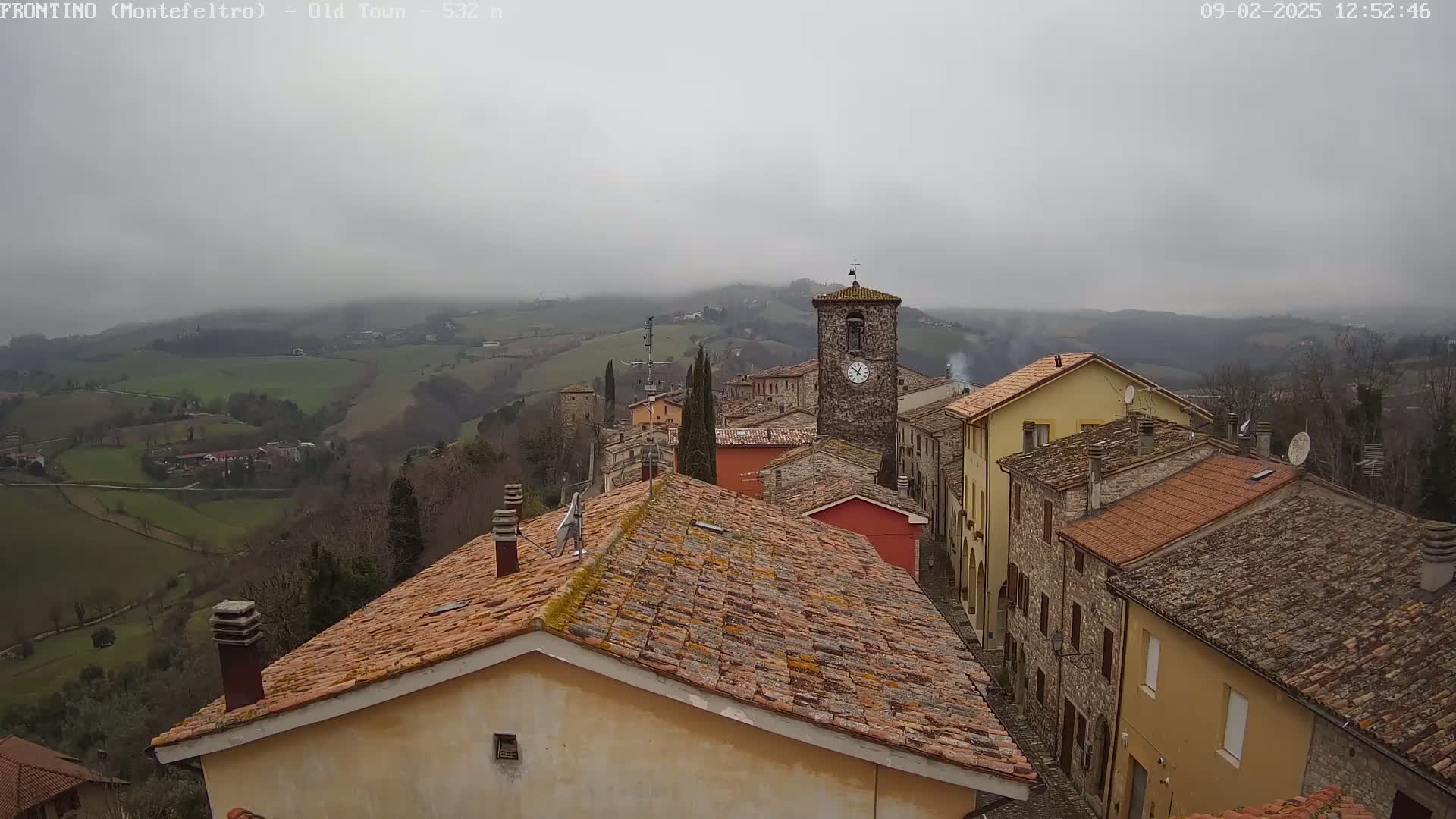 A view of a small town nestled in a hilly landscape on an overcast day, showing terracotta rooftops and a clock tower.