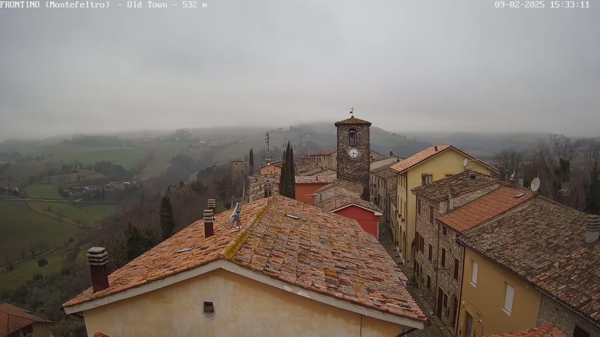 A foggy view of a small town nestled in a hilly landscape, with red-tiled rooftops and a clock tower visible among the buildings.