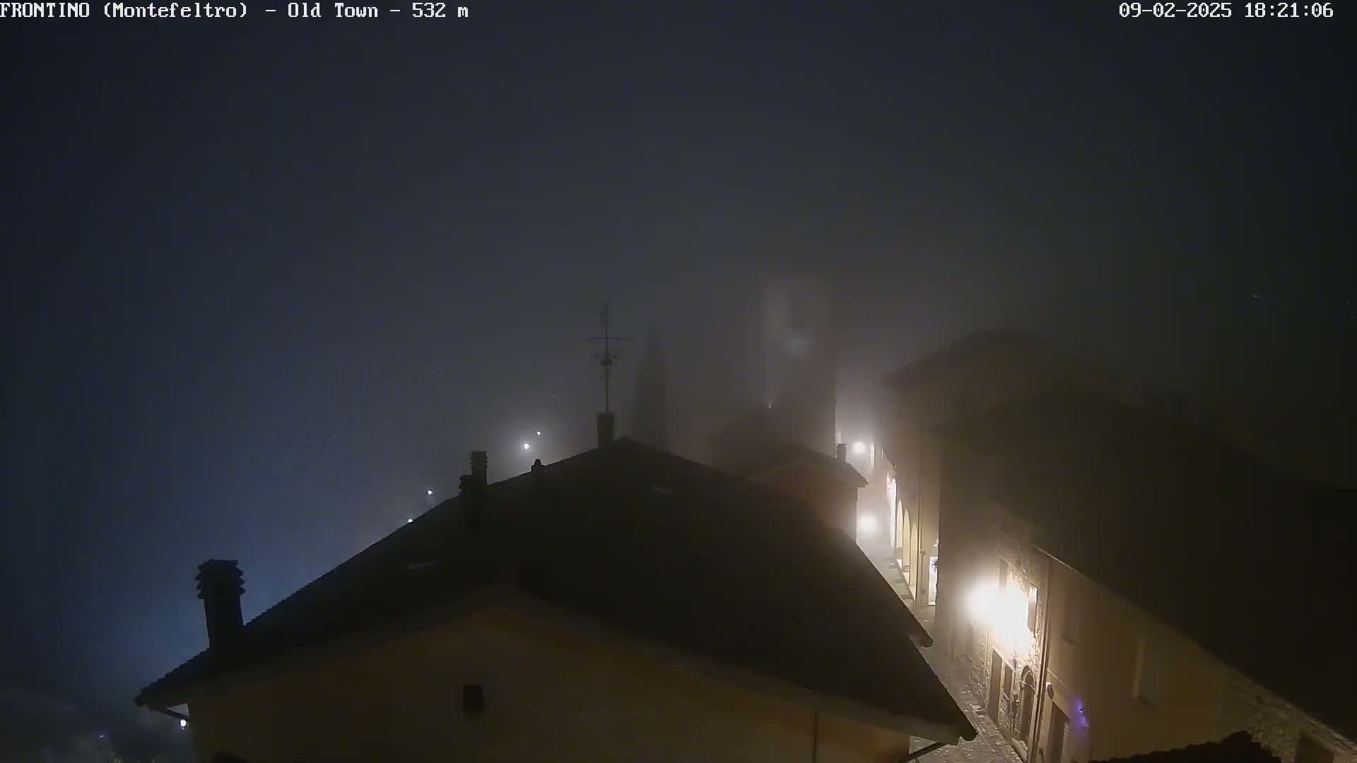A foggy night scene shows the rooftops and a dimly lit street of a town.