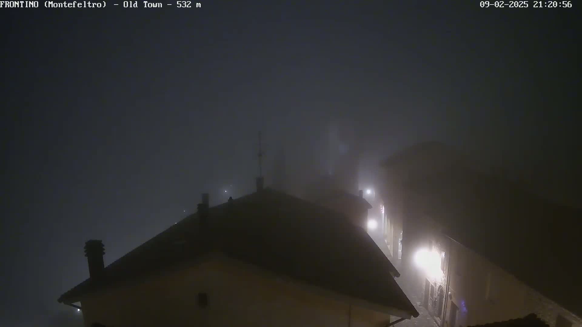 A foggy night scene shows the rooftops of buildings in a town, with faint lights visible through the dense fog.