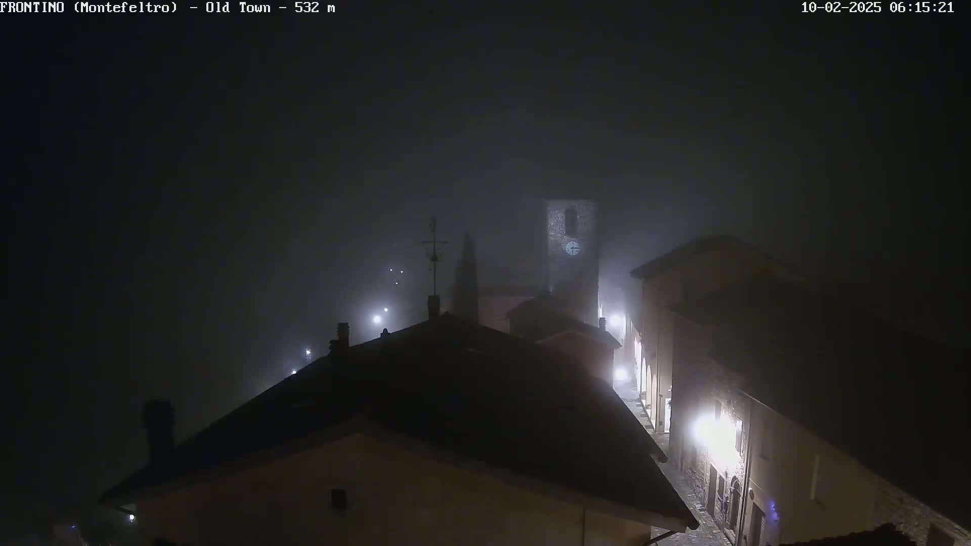 A foggy, night-time view of a dimly lit town square with several buildings, including a tall tower with a clock, partially obscured by fog.