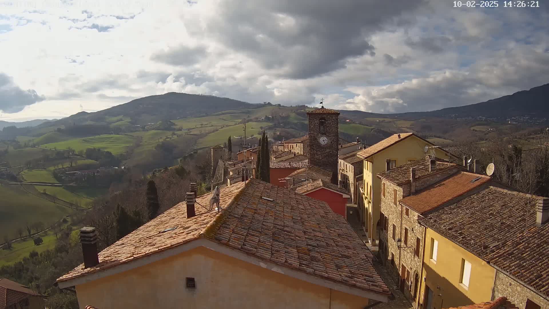 A quaint village with terracotta rooftops nestles in a valley, overlooked by rolling green hills under a partly cloudy sky.