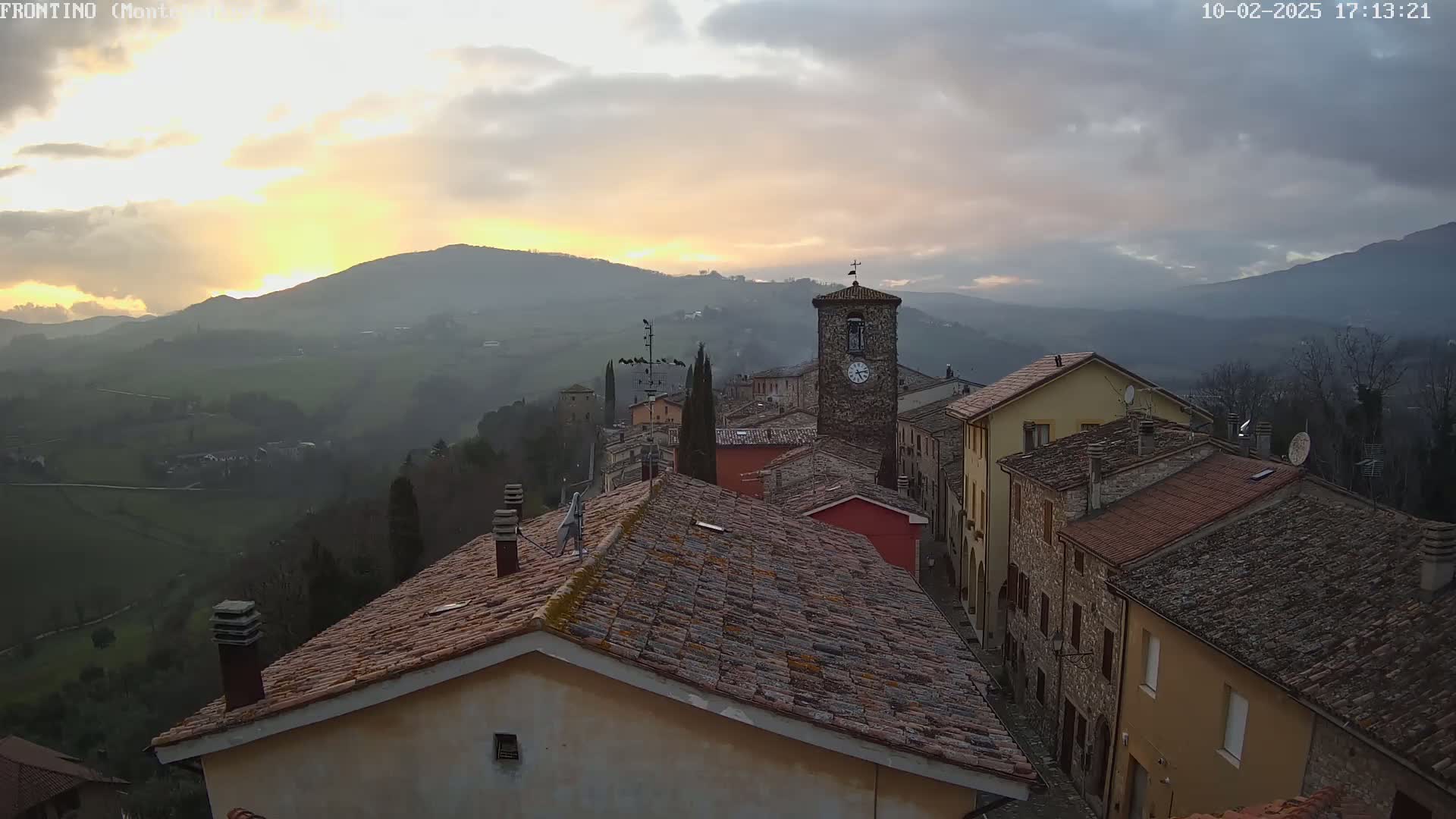 A hillside village with terracotta rooftops and a clock tower is seen at sunset under a cloudy sky.