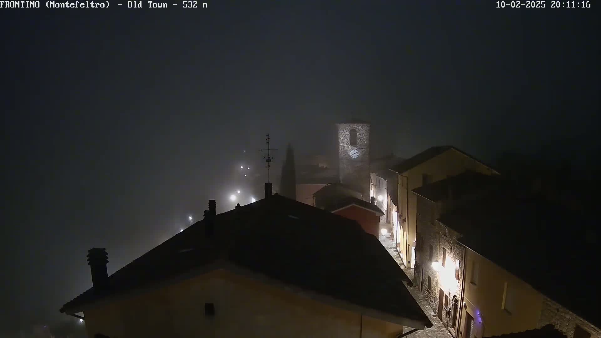 A fog-shrouded, nighttime view of a dimly lit old town, featuring stone buildings and a clock tower.