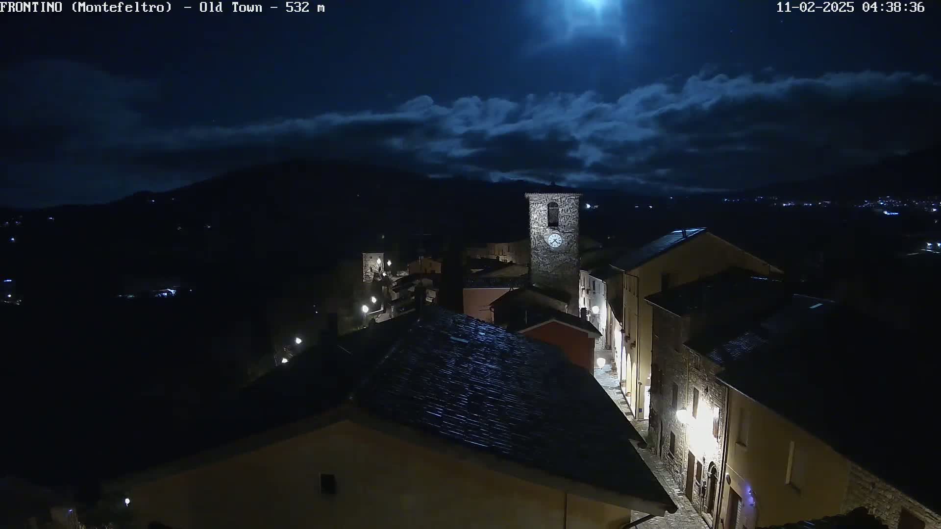 A nighttime, high-angle view of a small, dimly lit town nestled in a valley under a partly cloudy moonlit sky.