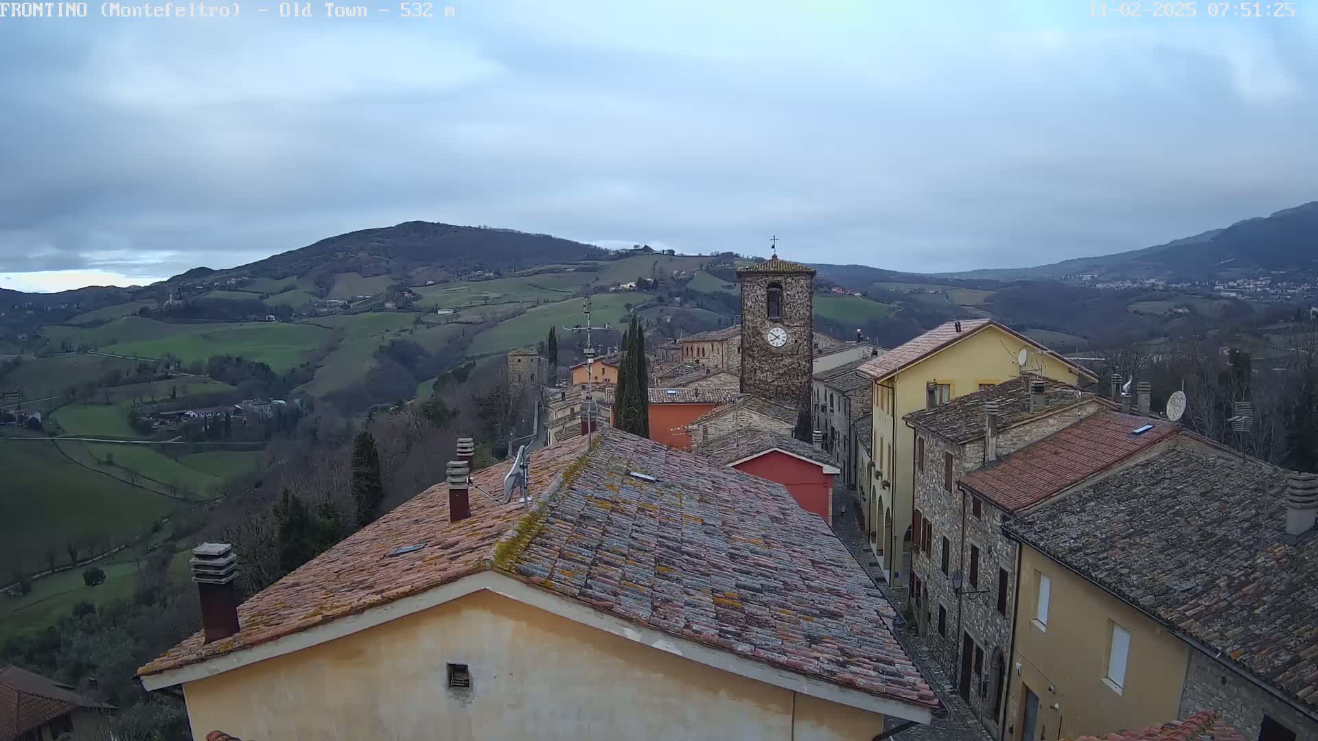 A view of a small town nestled in a hilly landscape under a cloudy sky, showing buildings with terracotta roofs and a prominent clock tower.