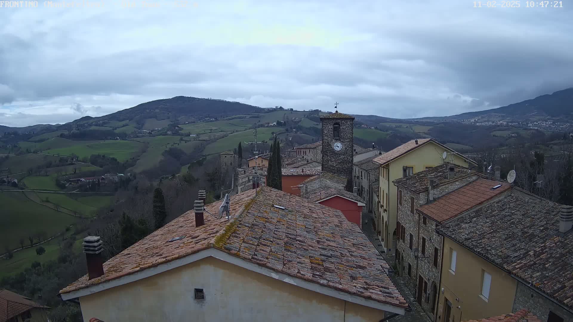 A view from a rooftop shows a small village nestled in a hilly, green landscape under a cloudy sky.