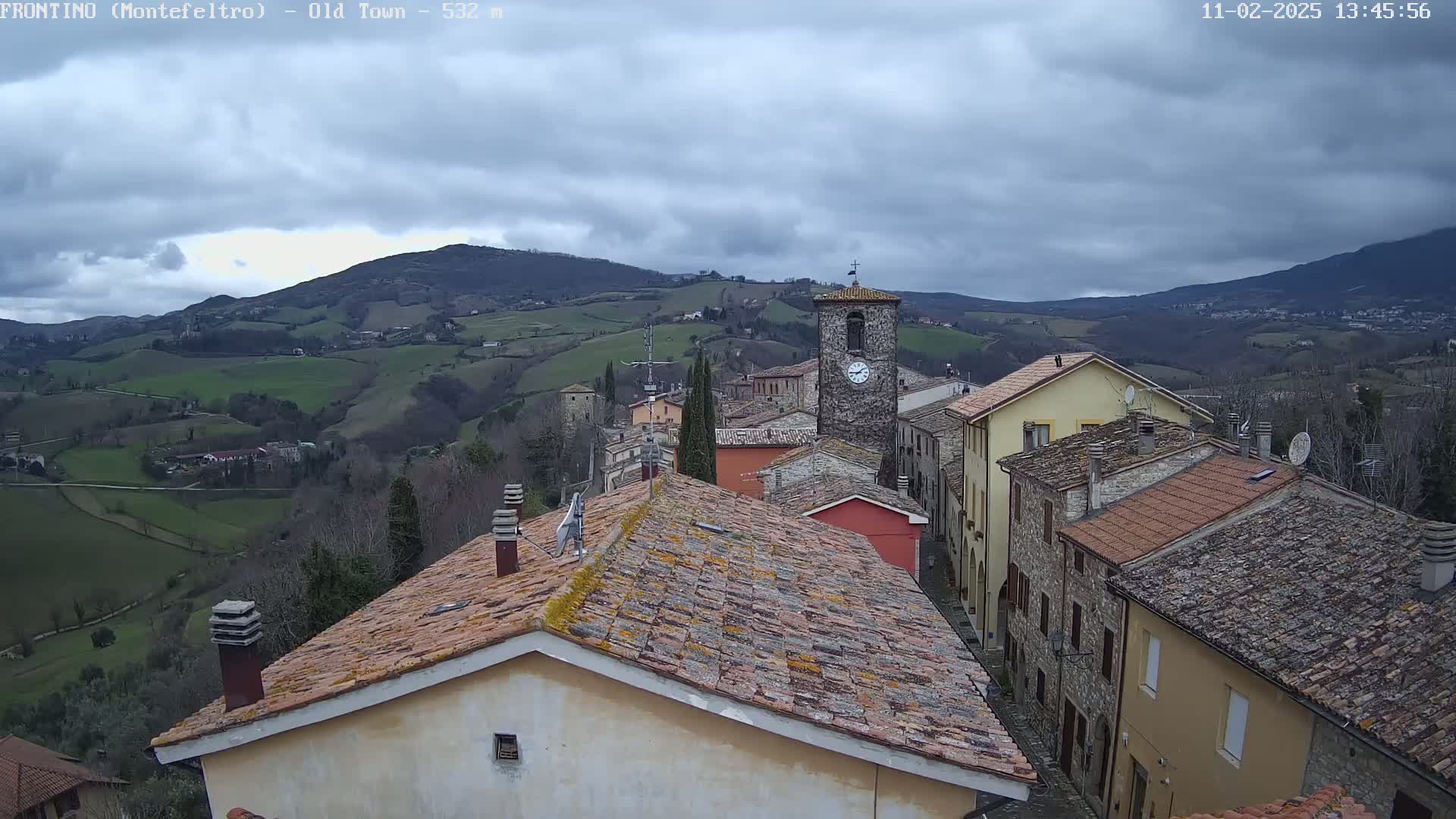 A view from a rooftop shows a small town nestled in a valley under a cloudy sky, with rolling hills and mountains in the distance.