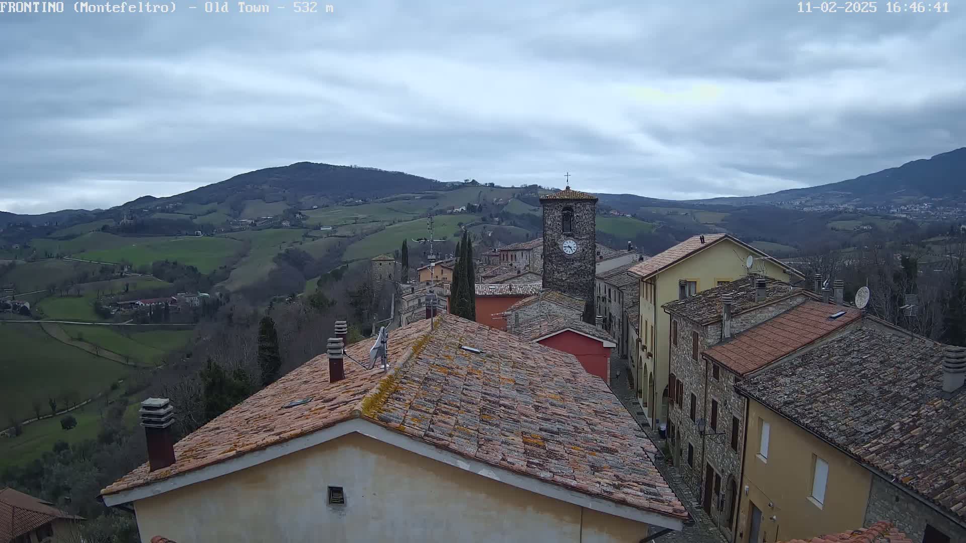 An overcast day reveals a quaint village nestled in rolling green hills, with a stone clock tower prominent among the terracotta rooftops.