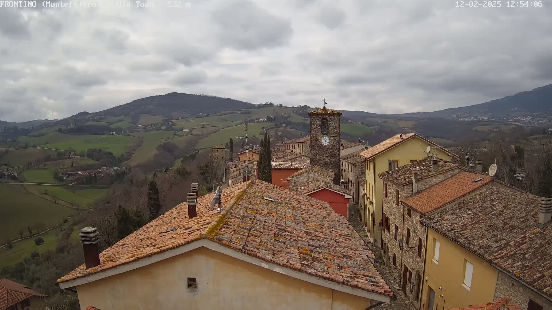 A small village nestled in a hilly landscape under a cloudy sky, with terracotta rooftops and a clock tower visible.