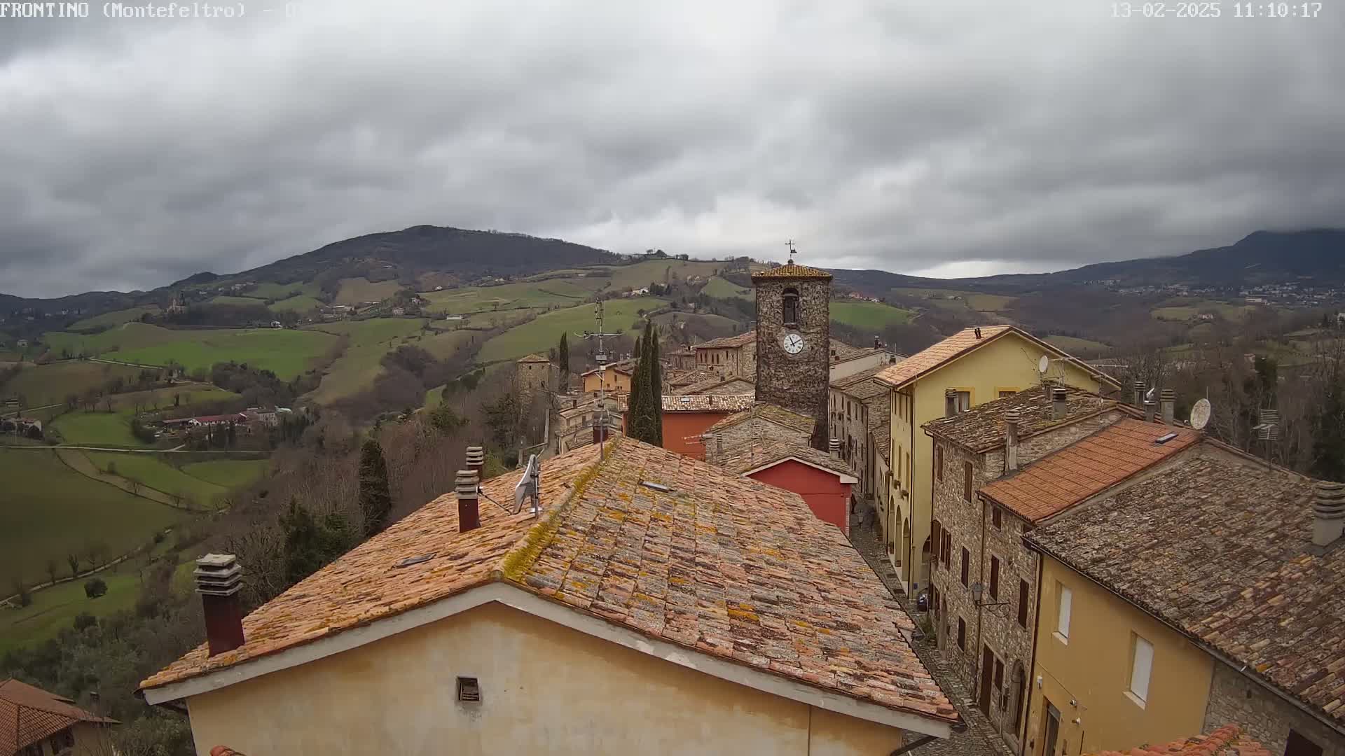 A small village of stone buildings with terracotta roofs nestles in a valley under a cloudy sky, overlooking rolling green hills.