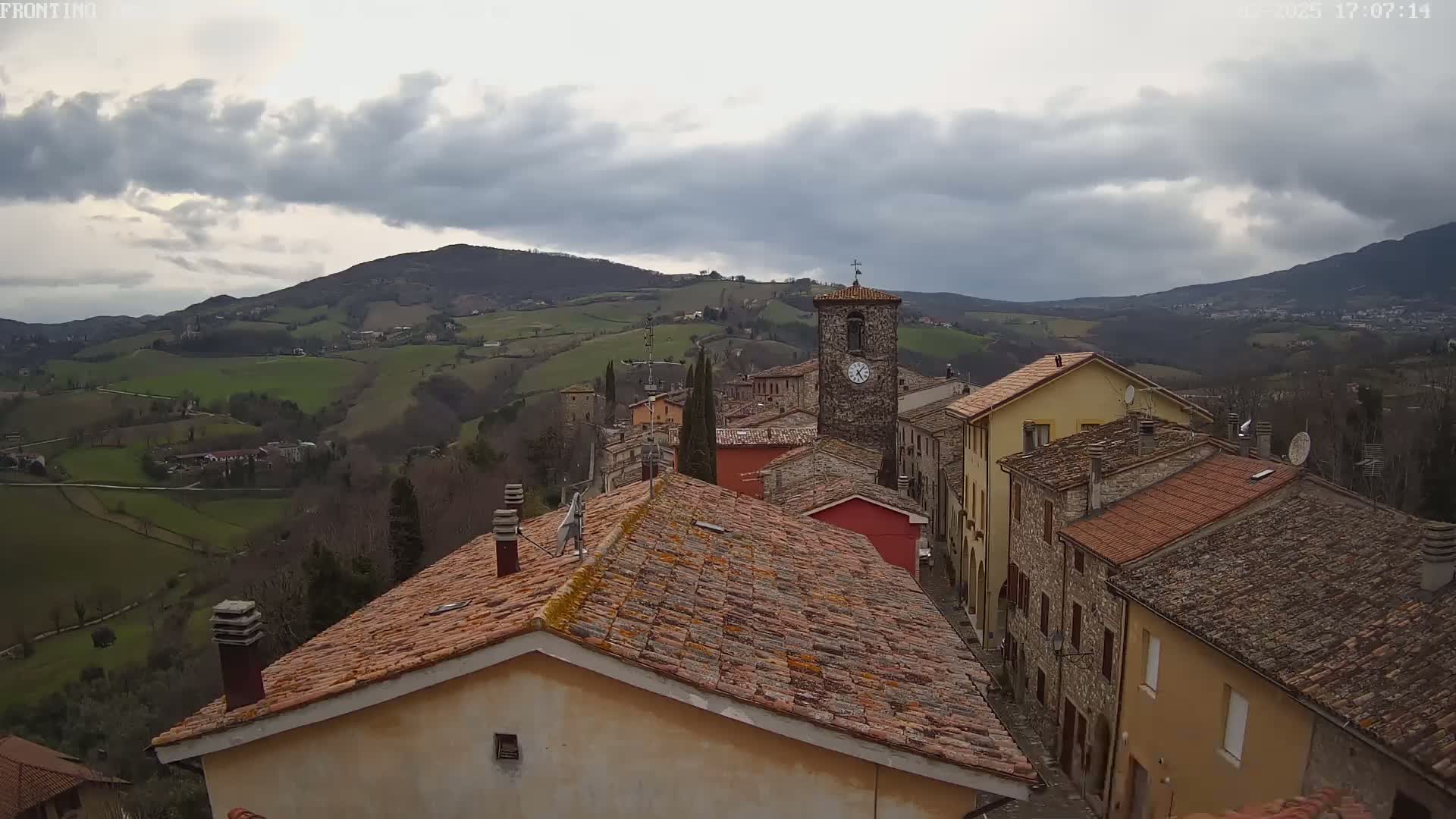 Overcast skies loom over a quaint village nestled in rolling green hills, with terracotta rooftops dominating the foreground.