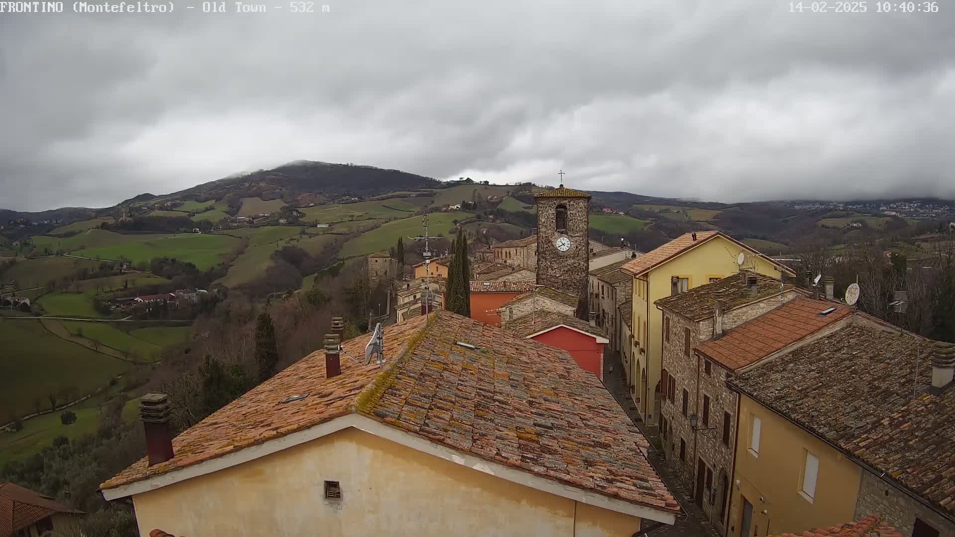 A small town nestled in rolling green hills under a cloudy sky is seen from a high vantage point, showing terracotta rooftops and a clock tower.