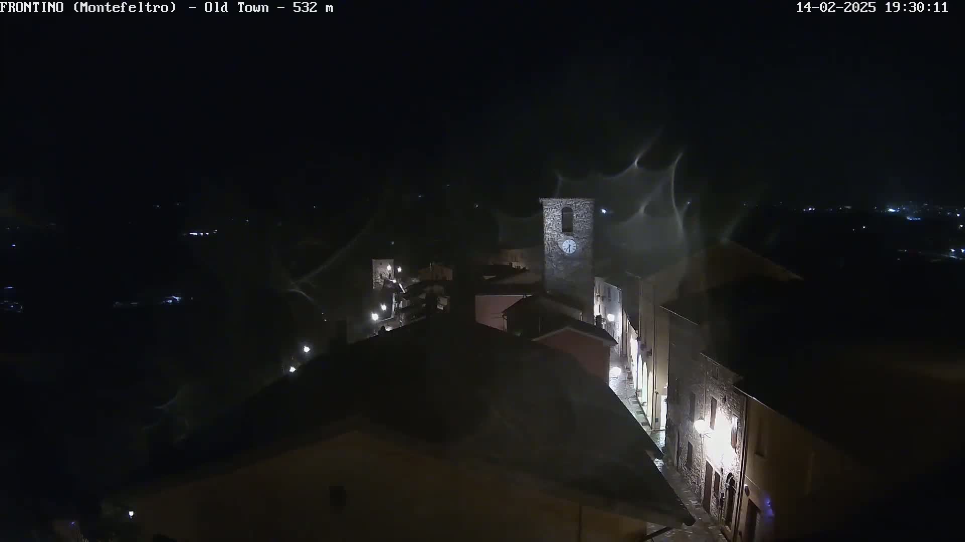 A nighttime, high-angle view of a small town with a clock tower, under windy conditions with what appears to be blowing debris or dust in the air.
