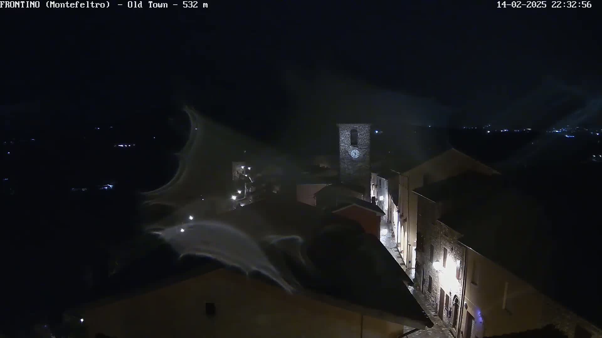 A nighttime, aerial view of a small town with a clock tower, illuminated by streetlights under a dark, possibly foggy sky.