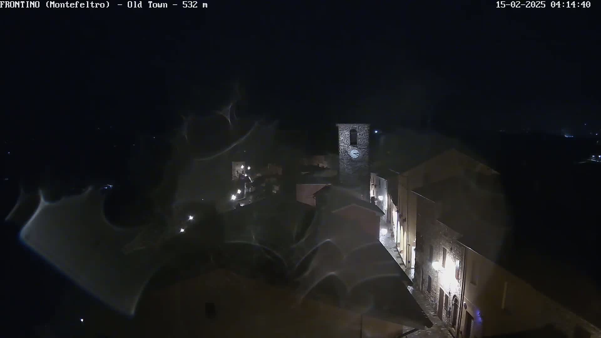 A nighttime, high-angle view of a small town, possibly in Europe, with dimly lit buildings and a prominent clock tower, under a dark, possibly cloudy sky.