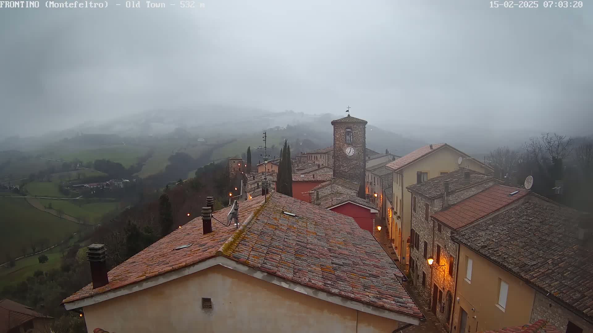 A misty, overcast view shows a quaint stone village nestled in a valley, with rooftops and a clock tower visible.