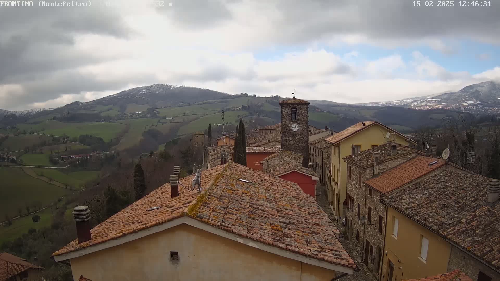 A stone village with terracotta roofs sits on a hillside overlooking snow-dusted mountains under a cloudy sky.