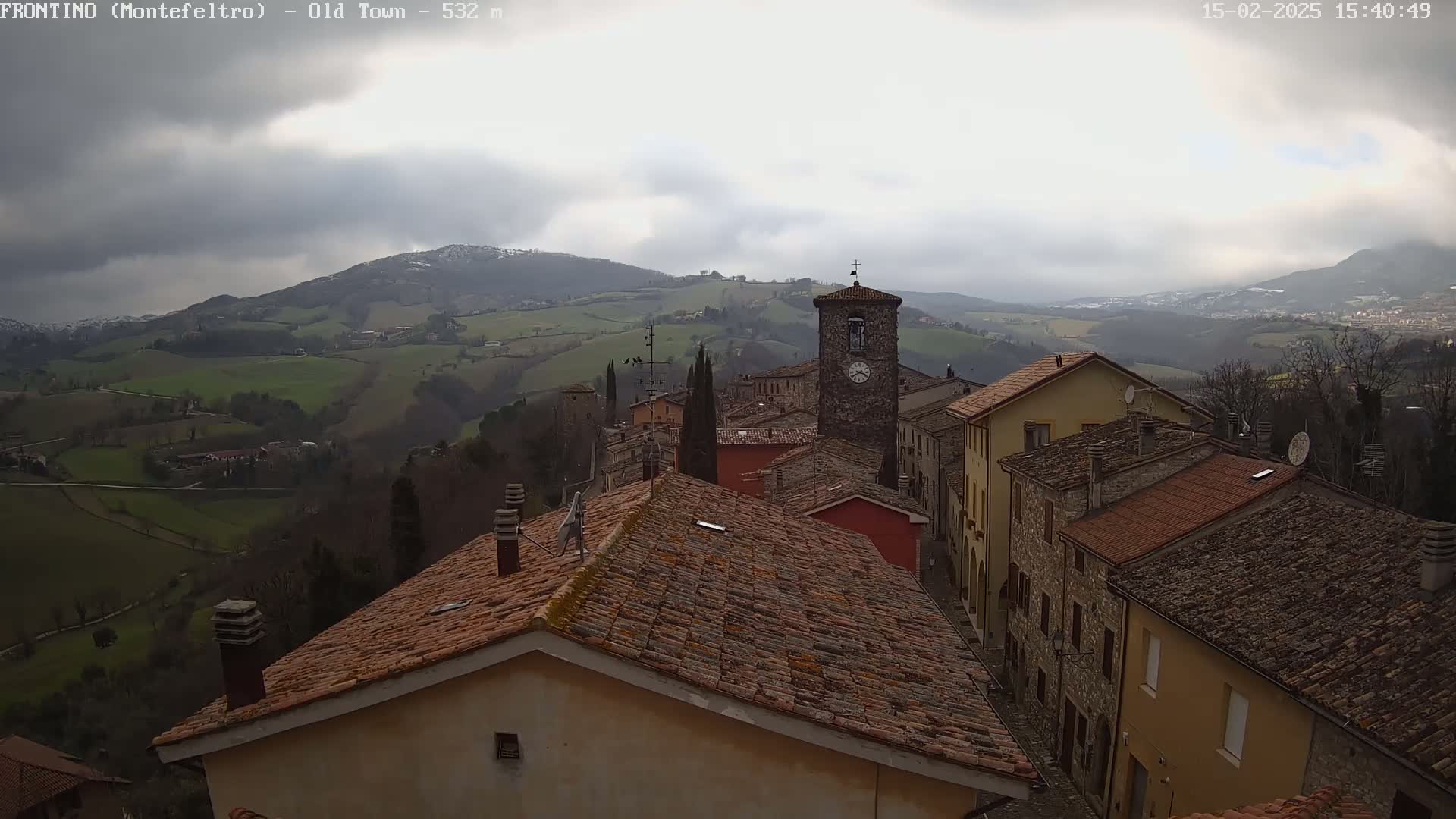 A stone village with red-tiled roofs sits on a hillside overlooking a valley under a cloudy sky.