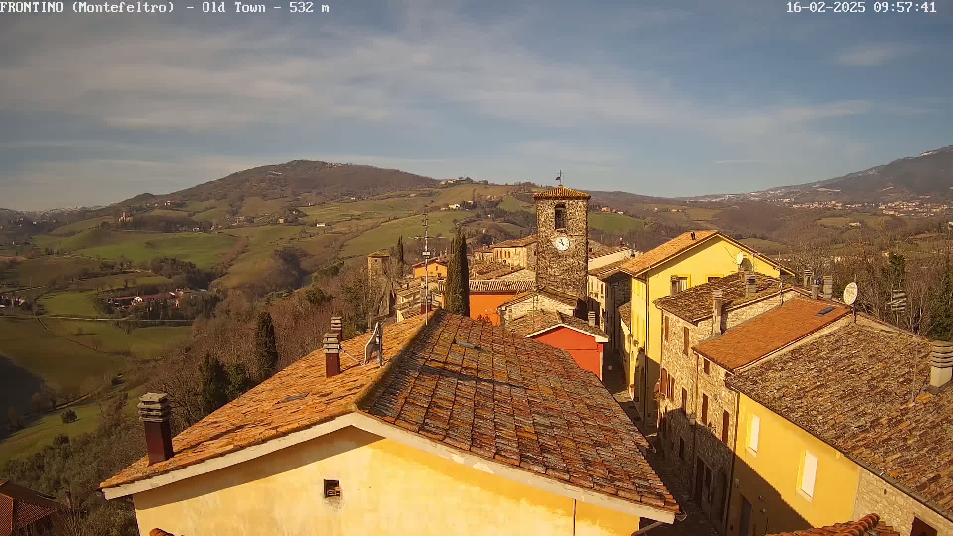 A sunny day reveals a quaint village nestled in rolling hills, with terracotta rooftops and a prominent clock tower.