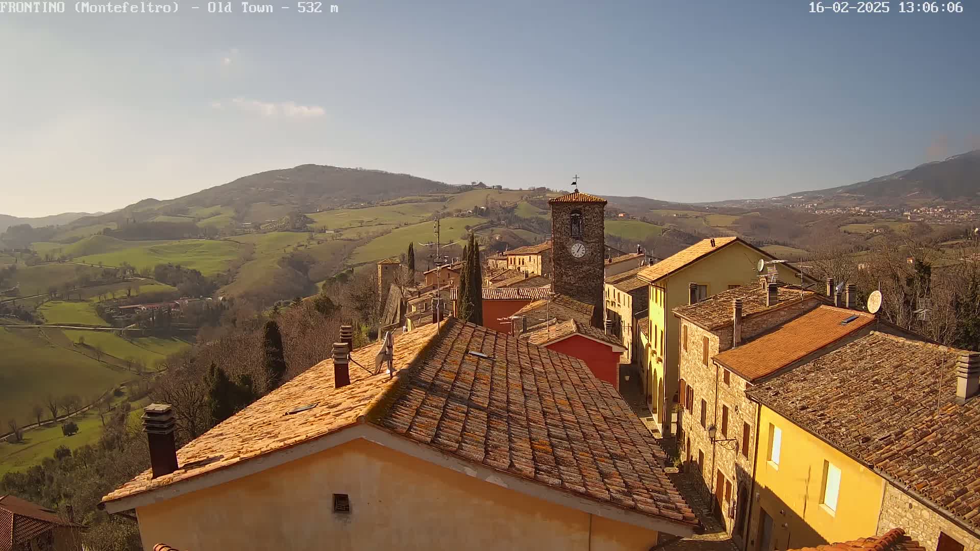 A sunny day reveals a small town nestled in a valley, with terracotta-roofed buildings and a clock tower, overlooking rolling green hills.