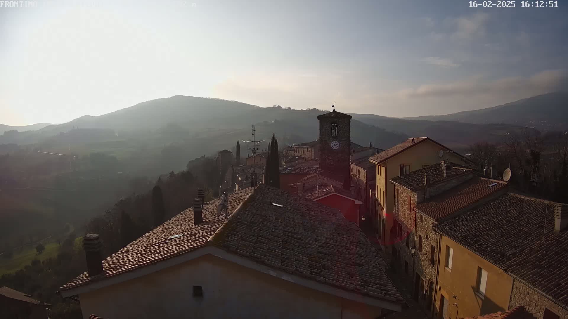 A hazy, sunlit view of a small village nestled in a valley between hills, showcasing terracotta rooftops and a prominent clock tower.