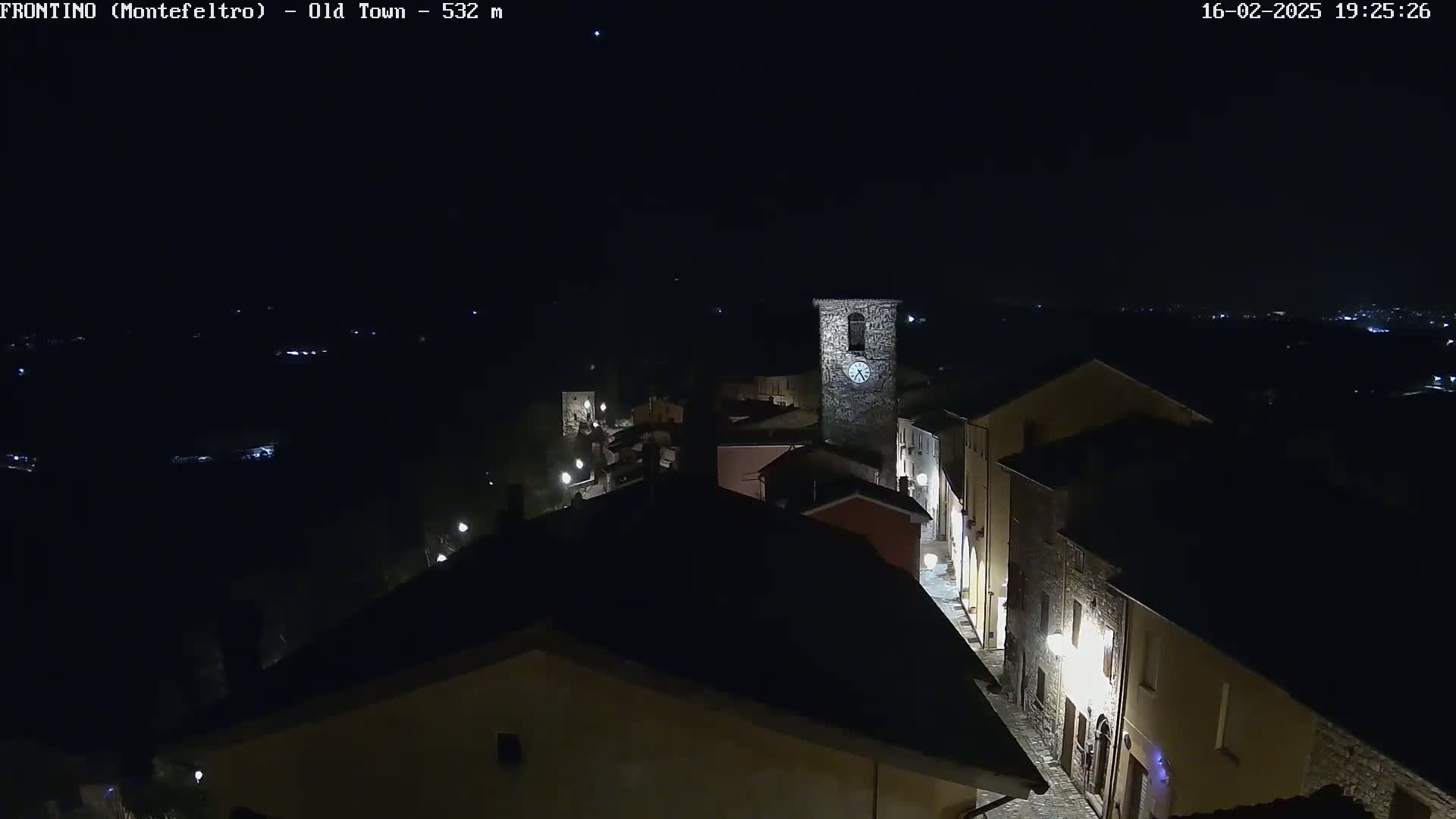 A nighttime, high-angle view shows a small town with dimly lit buildings and a clock tower, under a clear, dark sky.