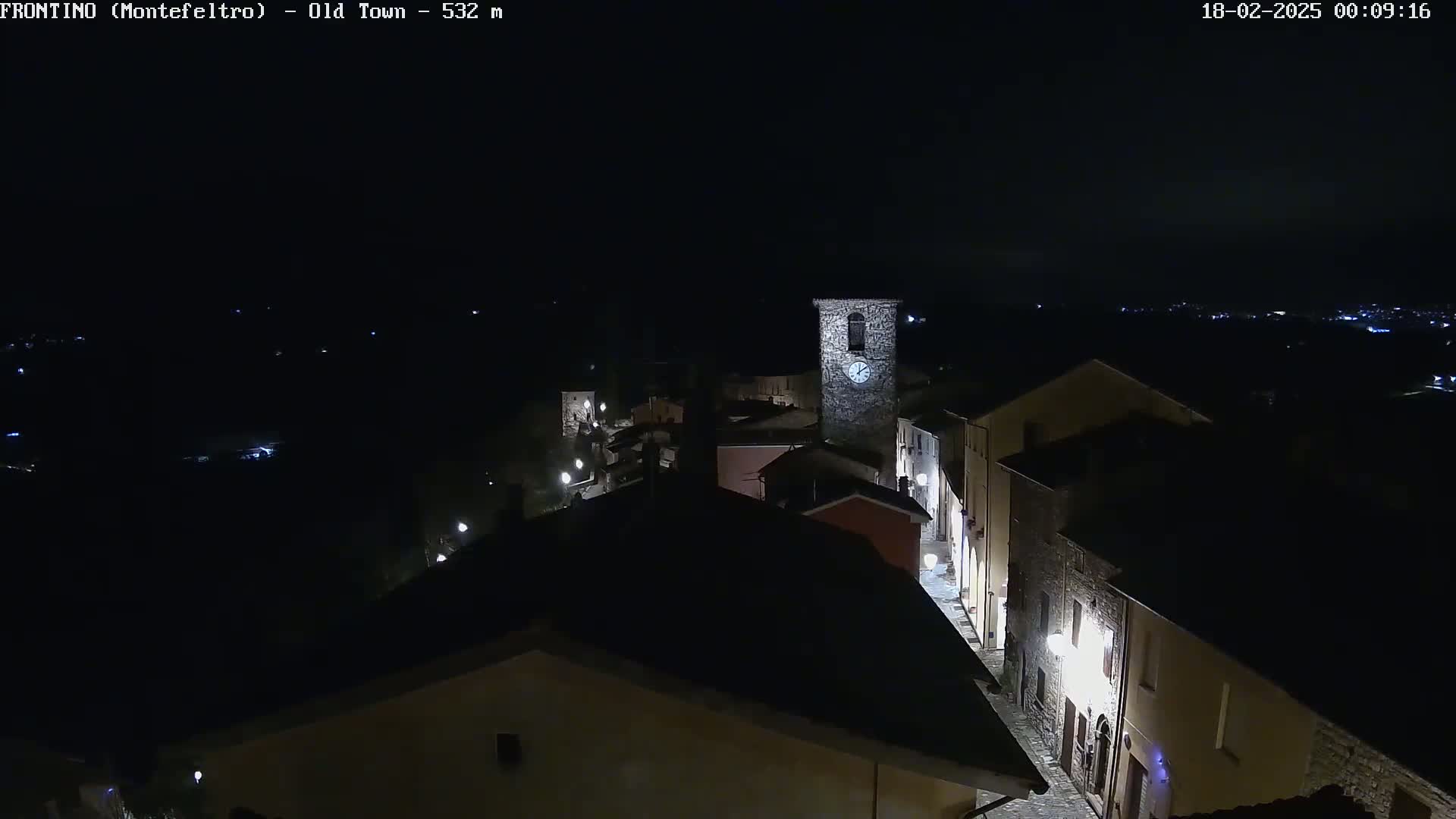 A nighttime, high-angle view of a stone village street illuminated by lampposts, featuring a clock tower and surrounding buildings.