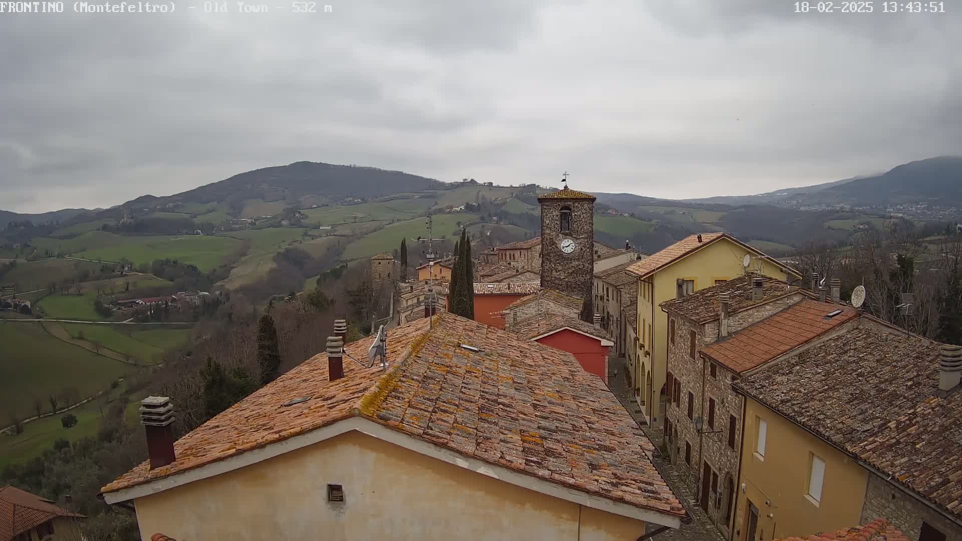 Overcast, a view of a stone village nestled in a valley, with rolling green hills and mountains in the background.