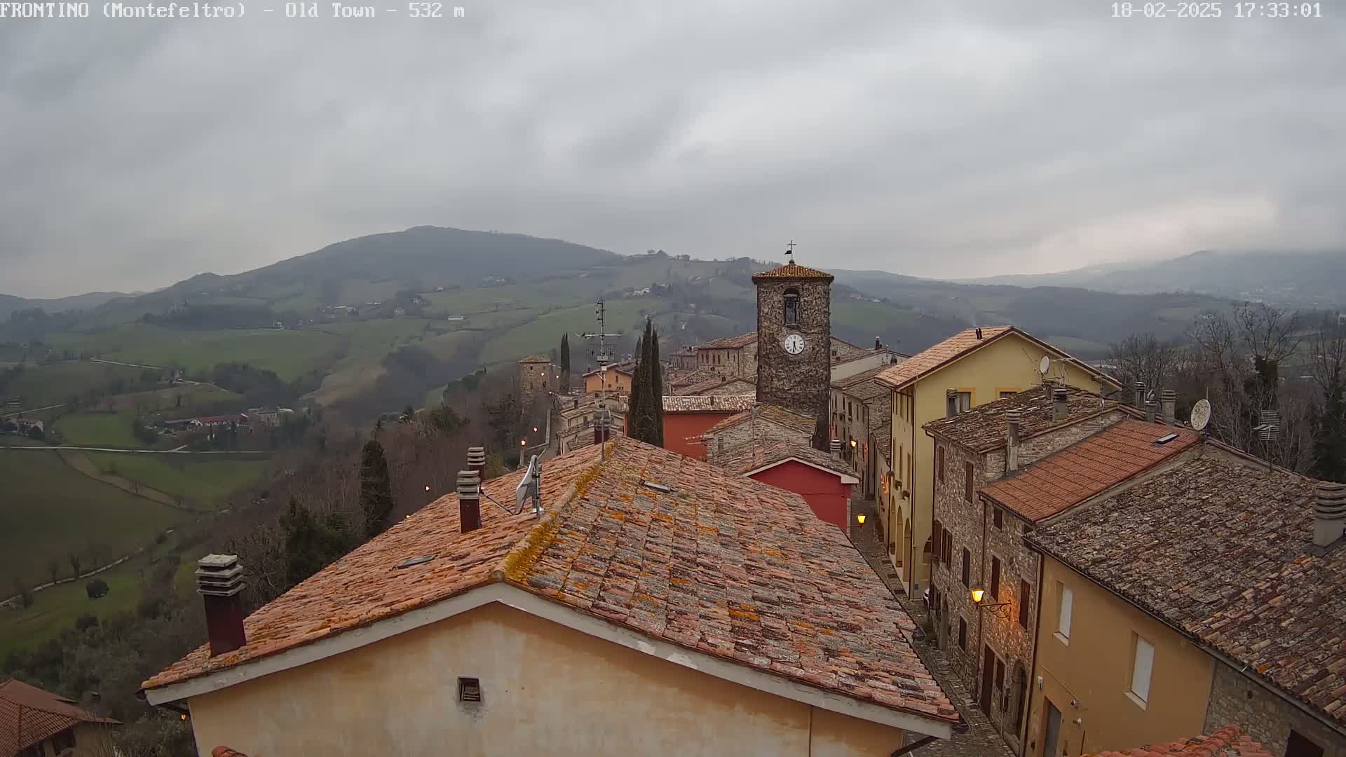 A view of a small town nestled in rolling hills on an overcast day, with terracotta roofs and a prominent clock tower visible.