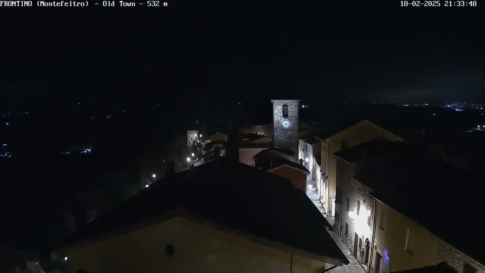 A nighttime aerial view shows a small, dimly lit town with stone buildings and a clock tower, under a clear, dark sky.