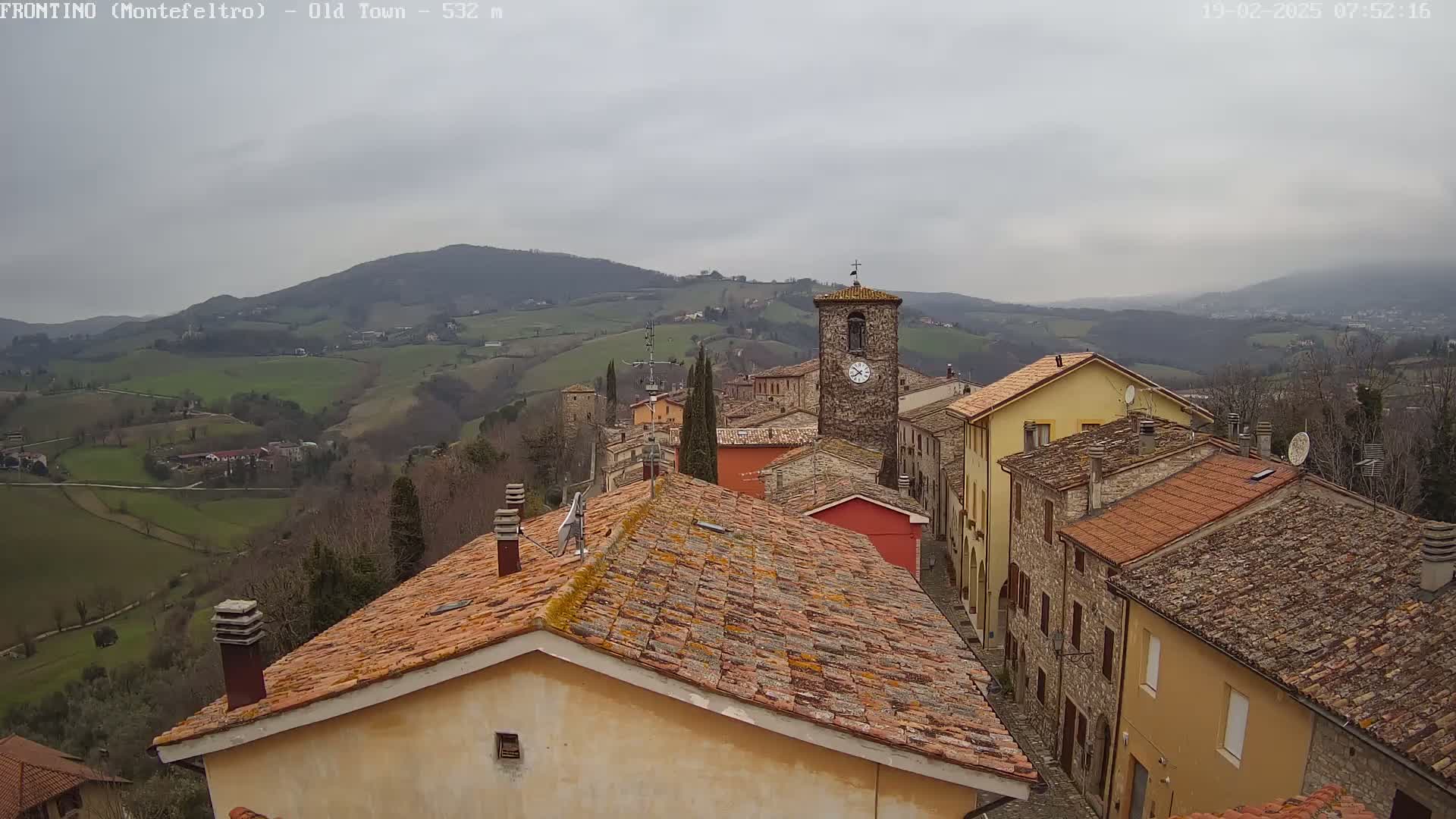 A stone village with red-tiled roofs sits on a hillside overlooking a valley under a cloudy sky.