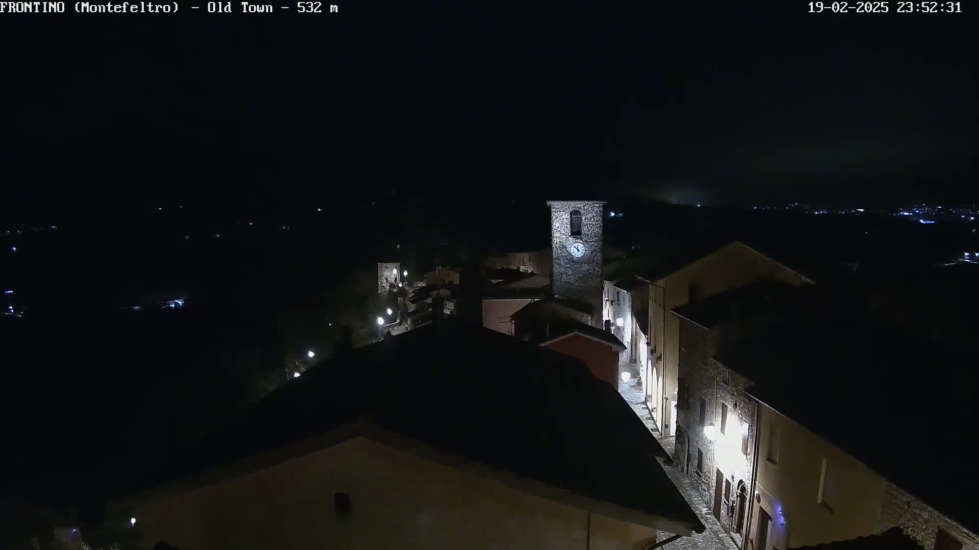 A nighttime, aerial view of a dimly lit, stone village with a clock tower, under a foggy sky.