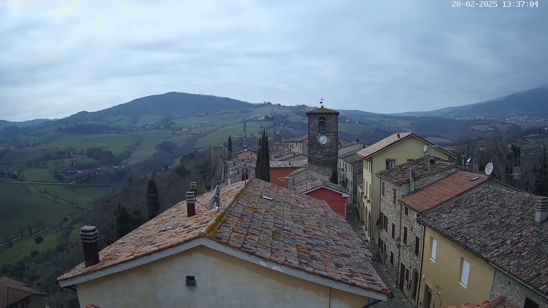 A stone village nestled in a hilly landscape under a cloudy sky overlooks rolling green fields and distant mountains.