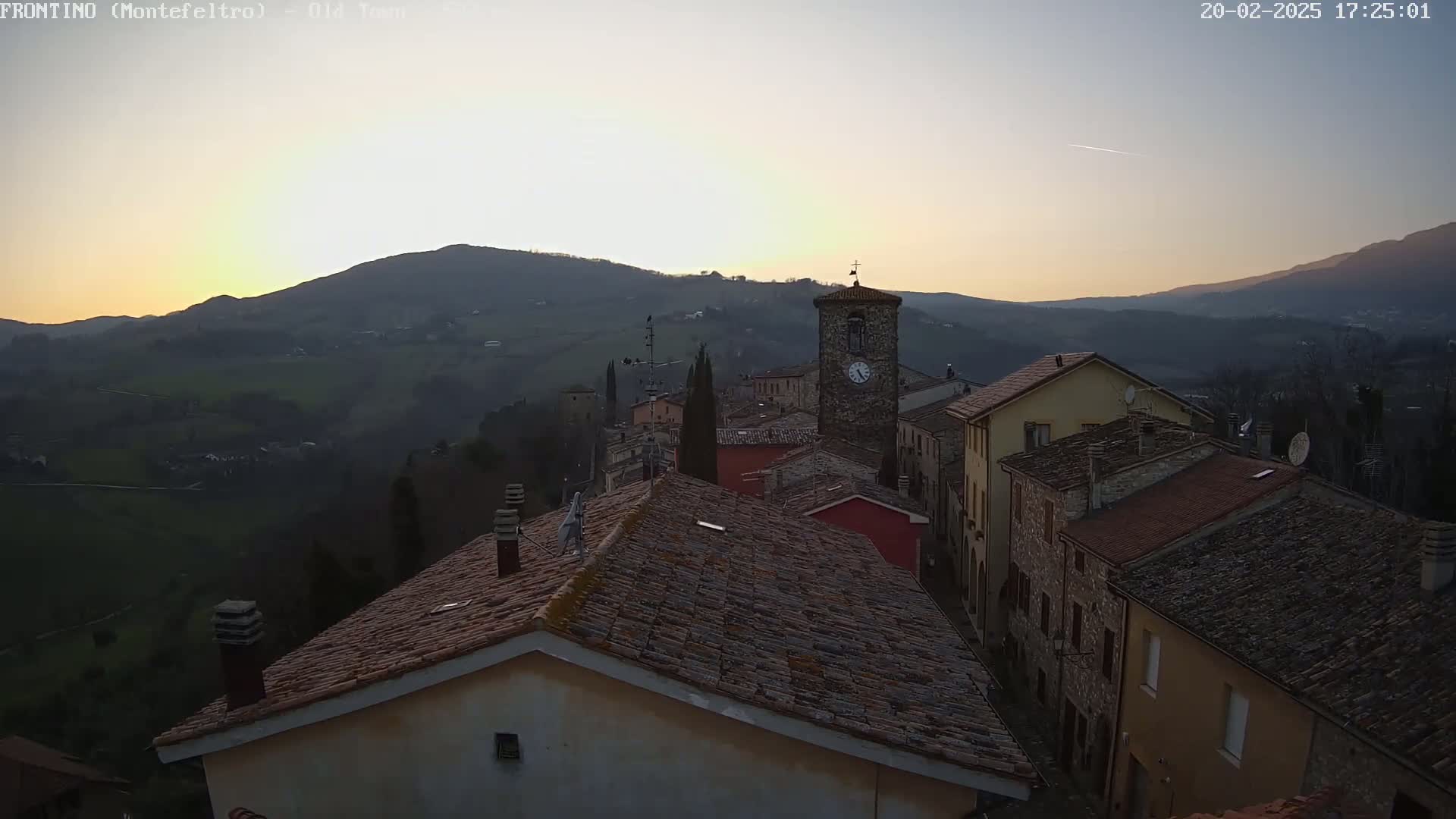 A hillside village with terracotta rooftops and a clock tower is seen at sunset under a clear sky.