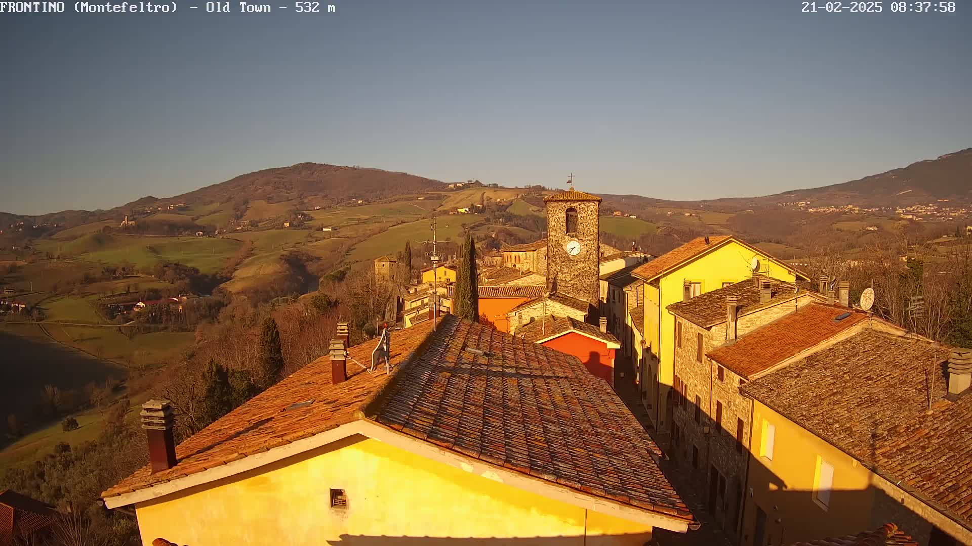 A sunlit view of a small town nestled in a valley, surrounded by rolling hills under a clear sky.