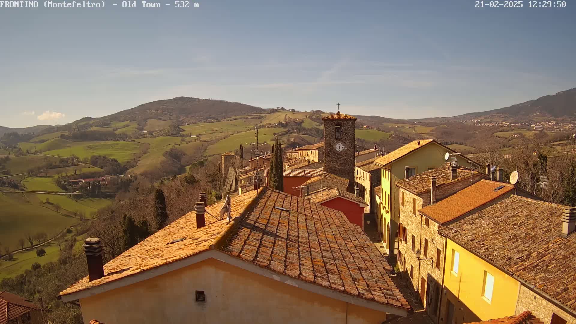 A quaint village nestled in a rolling hillside, under a clear sunny sky, features terracotta-tiled roofs and a prominent clock tower.
