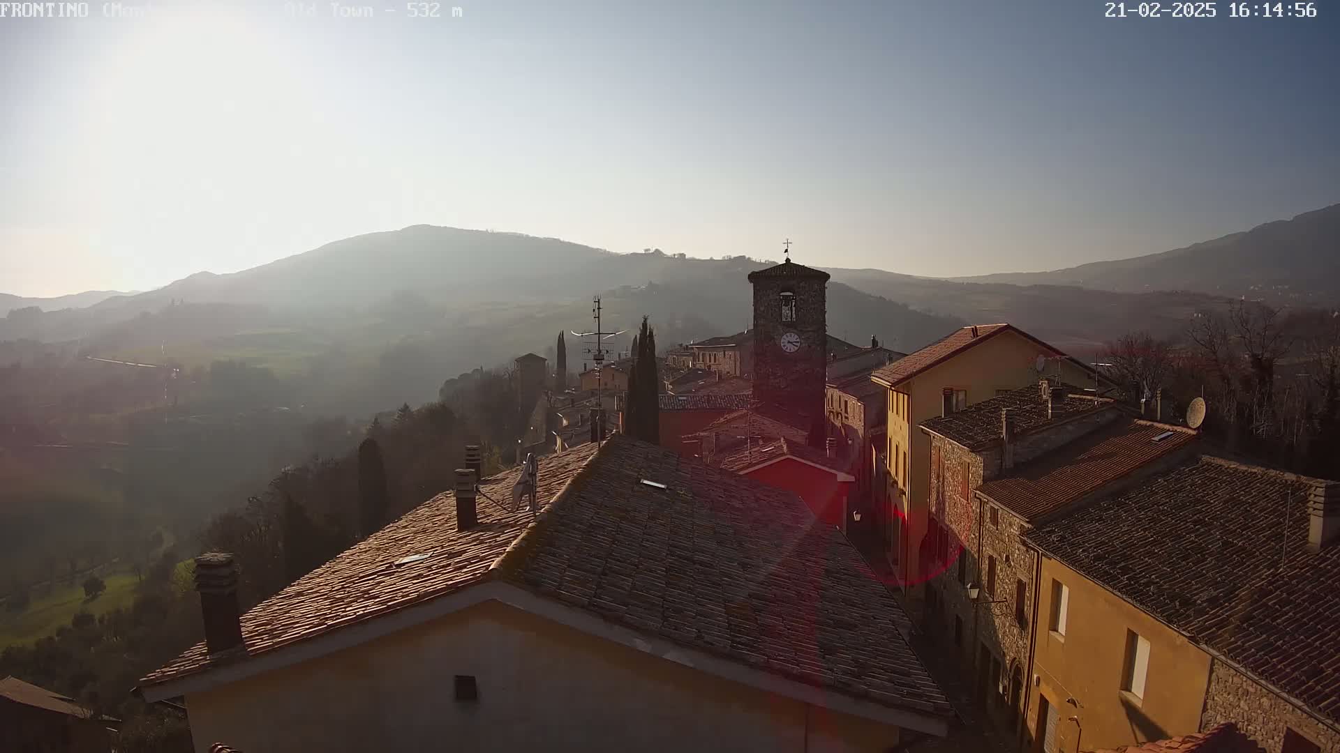 A hazy, sunlit view of a village nestled in a valley, with terracotta-roofed buildings and a clock tower, surrounded by rolling hills.