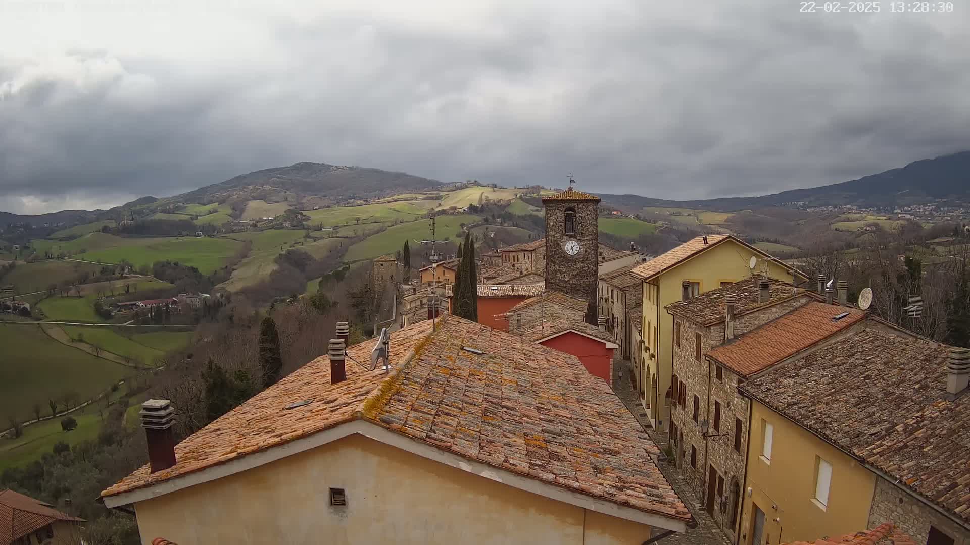 A small village with terracotta rooftops sits atop a hill overlooking a rolling green landscape under a cloudy sky.