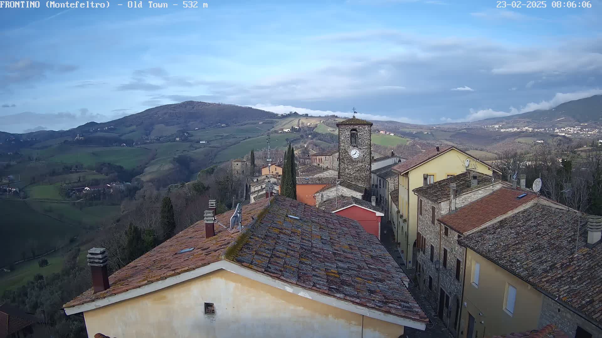 A stone village with a clock tower is nestled in a valley, overlooked by rolling hills under a partly cloudy sky.