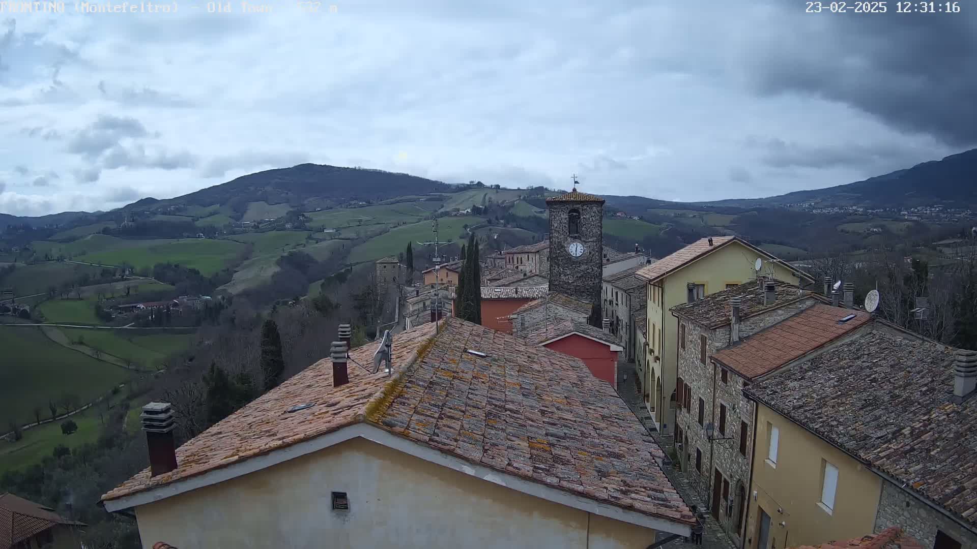 A small village nestled in a valley, under a cloudy sky, is seen from a rooftop perspective, showing tiled roofs, stone buildings, and a distant mountain range.