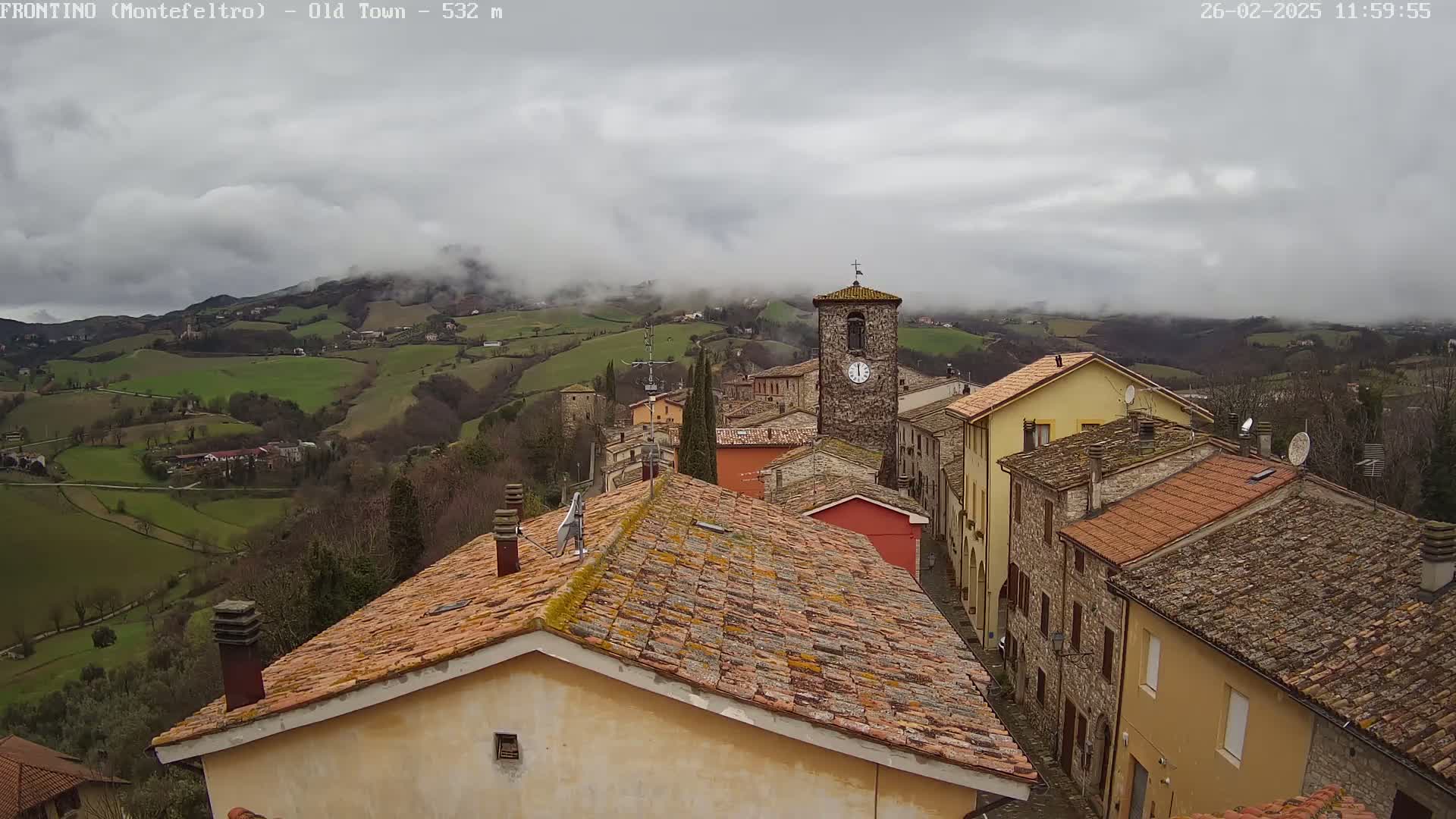 A stone village nestled in a valley, under a cloudy sky, overlooks rolling green hills partially obscured by fog.