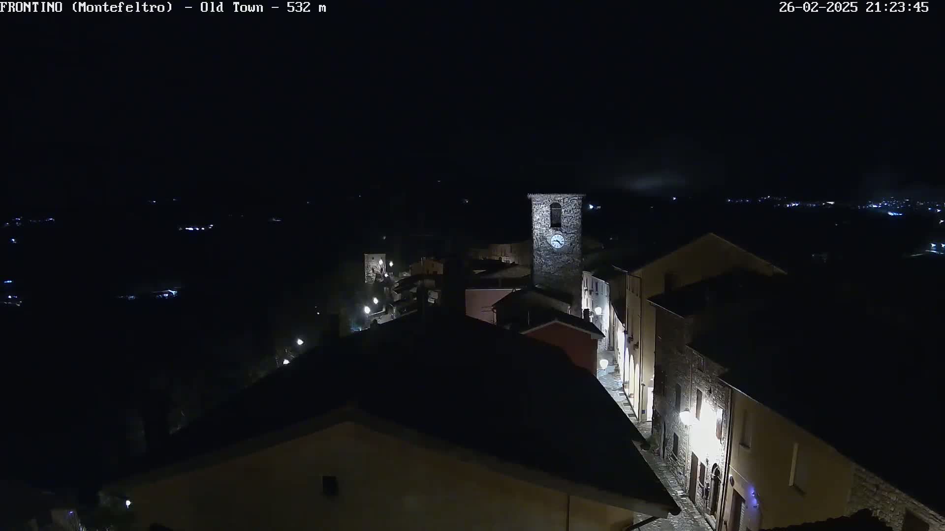 A nighttime, aerial view shows a dimly lit, stone village with a clock tower, under a dark, possibly cloudy sky.