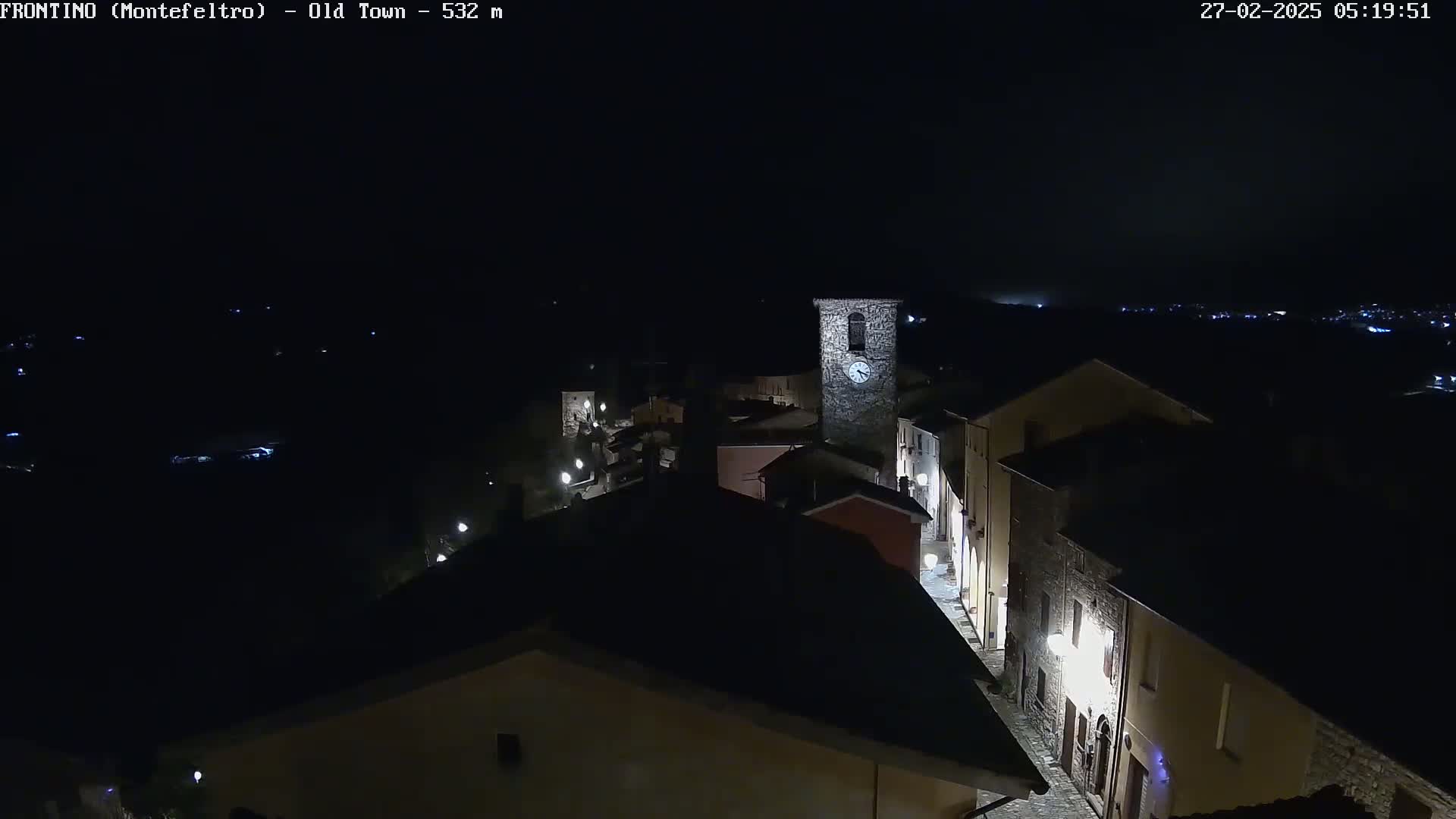 A nighttime, aerial view shows a small, stone village with a clock tower illuminated against a dark sky.