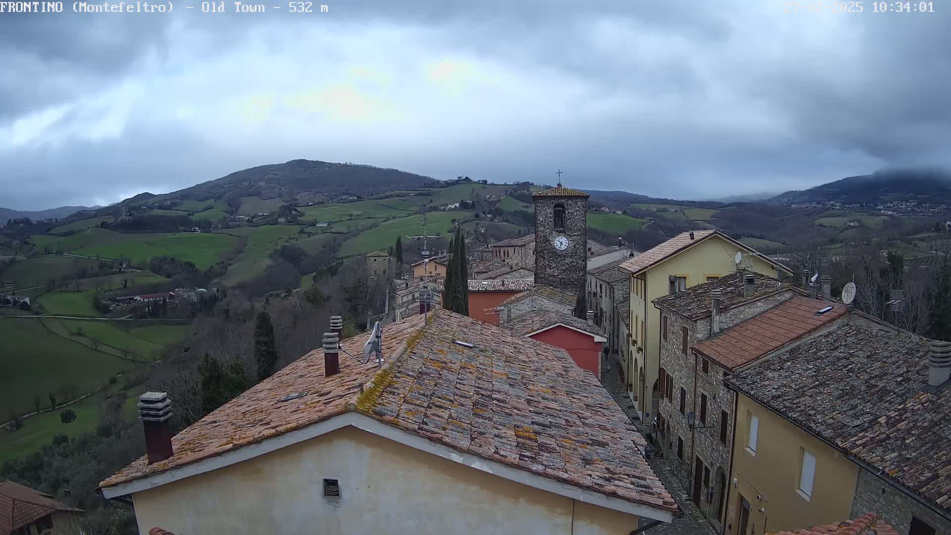A stone village with red-tiled roofs nestled in a valley, overlooked by rolling green hills under a cloudy sky.