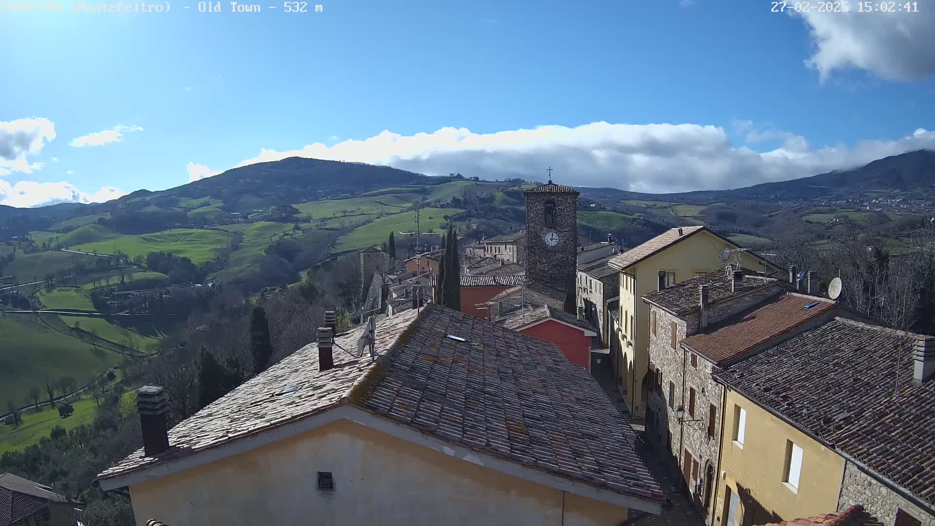 A small town nestled in a valley, under a partly cloudy blue sky, is seen from an elevated vantage point overlooking terracotta rooftops and rolling green hills.