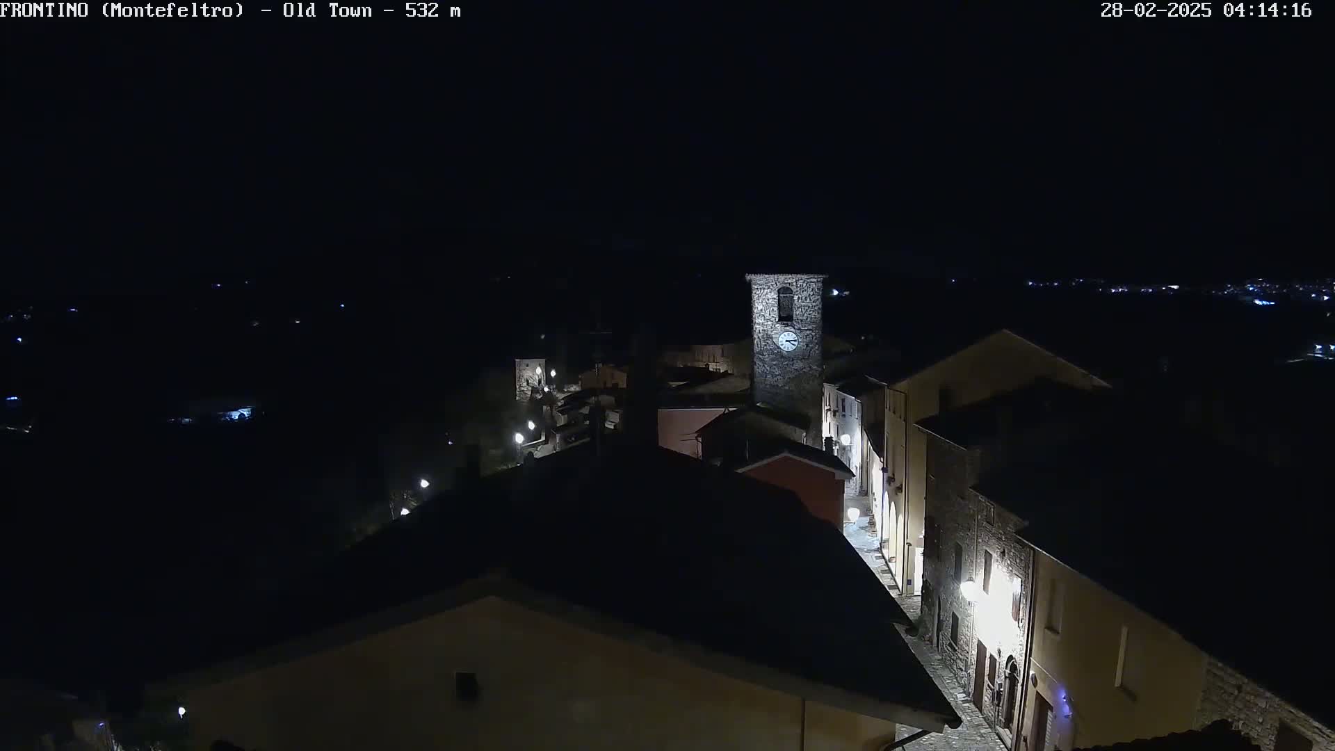 A nighttime, aerial view shows a small, dimly lit town with a clock tower, under a clear, dark sky.