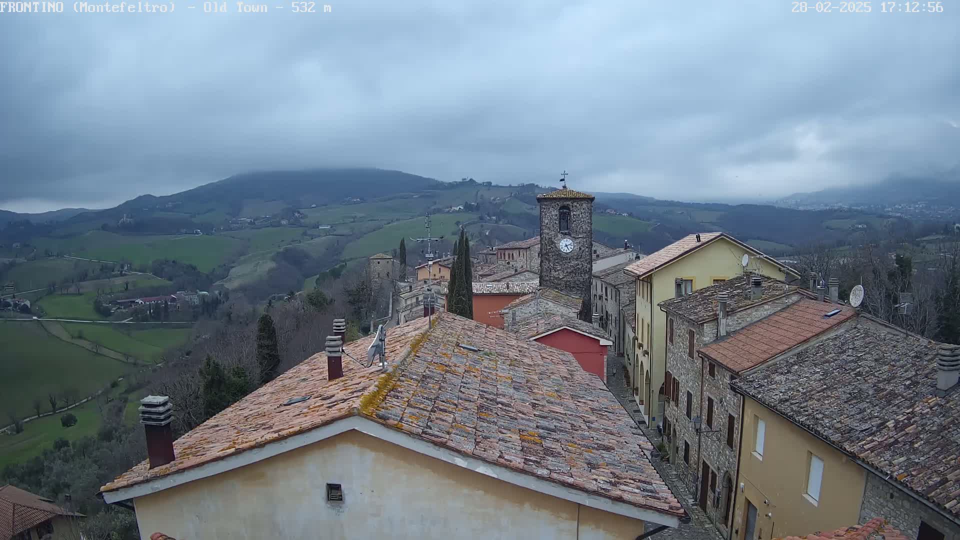 An overcast day reveals a quaint stone village nestled in rolling green hills, with a clock tower prominently featured in the center.