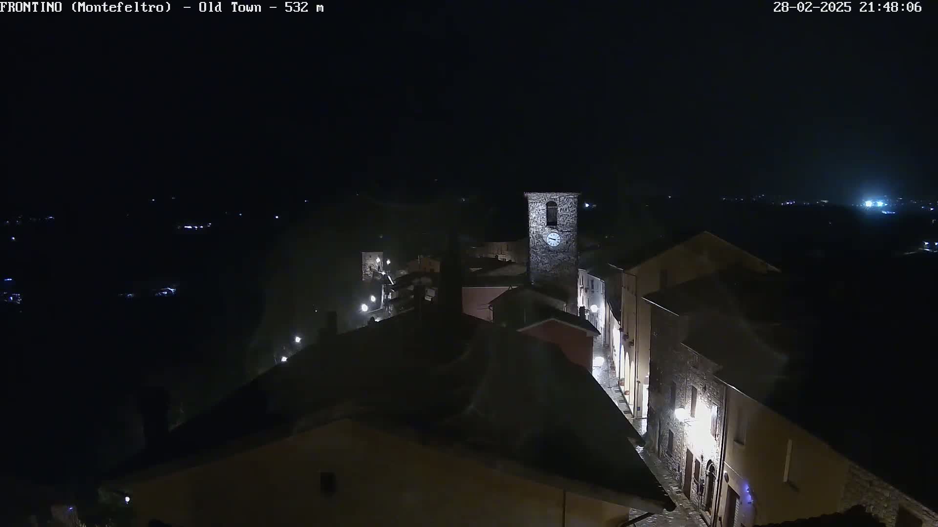 A nighttime, high-angle view of a small town's old town, illuminated by streetlights, with a clock tower visible.