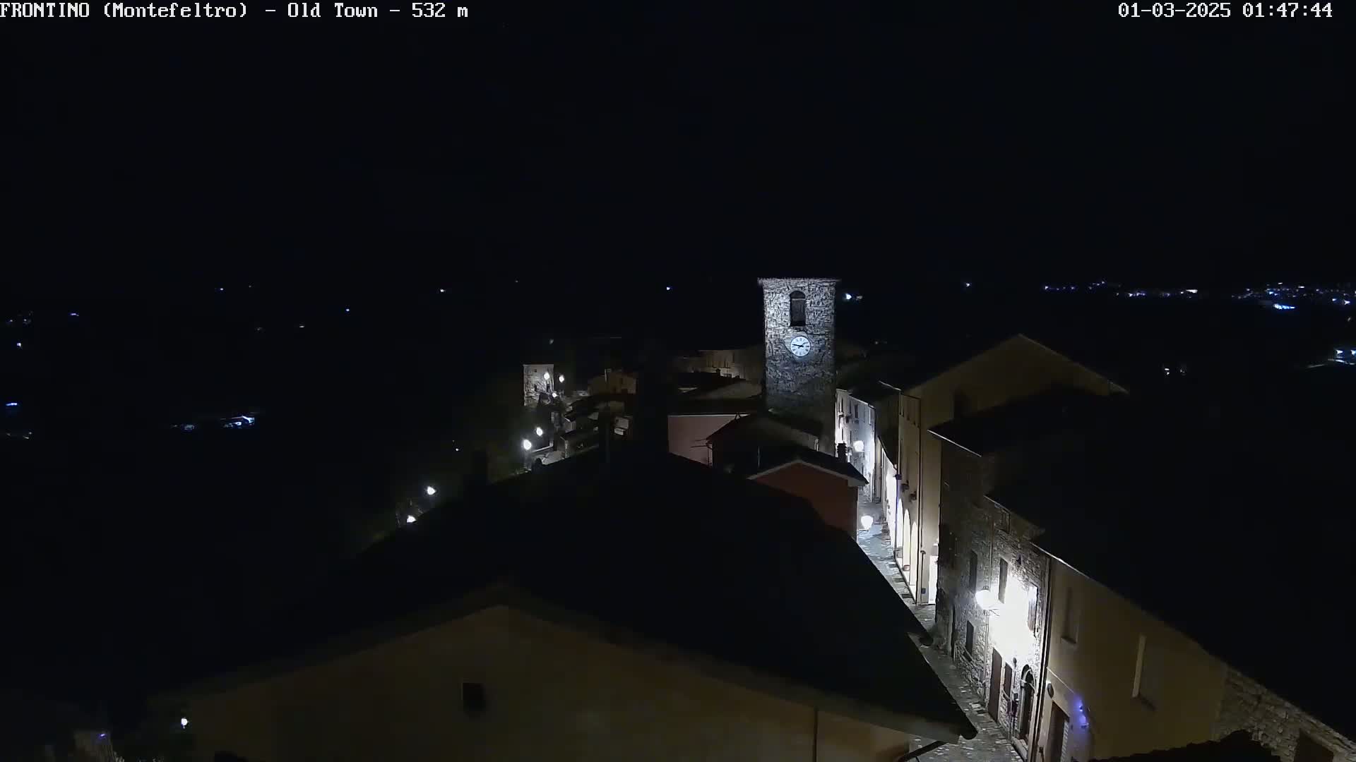 A nighttime, high-angle view of a small town with dimly lit streets and buildings, including a stone clock tower.